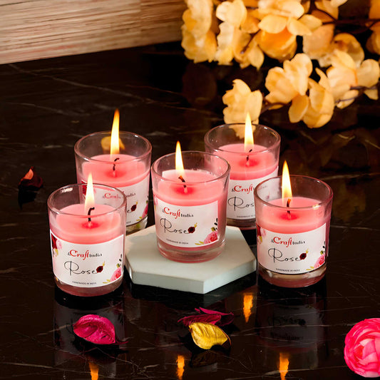 Set of five pink candles in glass holders with visible flames on a dark surface, surrounded by decorative flowers.